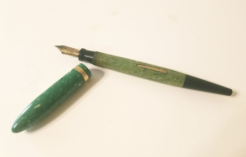 May include: A green and black fountain pen with a gold band. The pen is closed and the cap is off. The pen is on a white surface.
