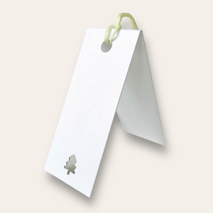 May include: A folded white rectangular paper tag with a small die-cut evergreen tree shape at the bottom. A circular hole is punched near the top fold, threaded with a pale yellow string loop for attachment to items or gifts.