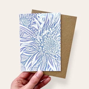 May include: A rectangular greeting card featuring a light blue and white floral print with a textured, stippled effect. The card is layered on top of a plain, light brown kraft paper envelope, showcasing a high-contrast botanical illustration.