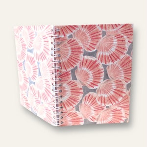 May include: A spiral-bound notebook featuring a repeating pattern of pink and white scallop shells against a grey background. The cover displays a dense arrangement of these shells, and the metal wire binding runs along the left side of the notebook.