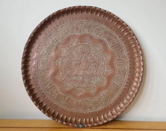 May include: A round copper tray with an intricate engraved design. The design features a central floral motif surrounded by Arabic script.
