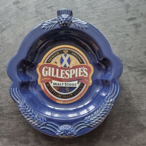 Op de afbeelding: Blauwe asbak van keramiek met een gouden en rood label met de tekst "Premium Original Gillespie's Malt Stout Scottish Malt Stout".