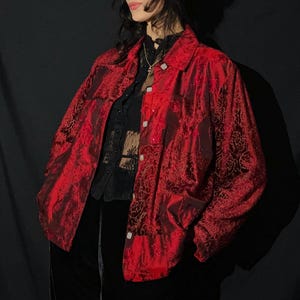 May include: A long-sleeved button-down jacket in a deep red hue featuring a tonal, intricate damask-style pattern throughout. The garment has a standard collar, a front button closure with light-colored buttons, and a structured, relaxed fit.