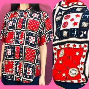 Puede incluir: Una camisa de manga corta con botones y temática náutica. La camisa presenta un diseño de patchwork con colores rojo, blanco y azul. El patrón incluye ancras, estrellas y diseños de cuerda.