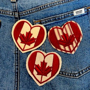 May include: Three red and white heart-shaped patches with a red maple leaf design. The patches are on a blue denim background.