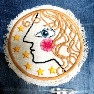 May include: Embroidered patch with a celestial design. The patch has a white background with a face, blue eye, and pink cheek. Gold stars and curly hair surround the face. The patch is bordered with gold thread and a fringe.