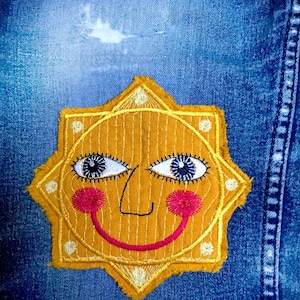 May include: A yellow embroidered sun with a smiling face, blue eyes, and pink cheeks, sewn onto a blue denim background.