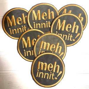 Peut inclure: Une collection d'écussons brodés circulaires présentant un fond gris foncé avec des coutures dorées. Chaque écusson affiche le texte "Meh innit." dans une police grasse sans empattement, avec une bordure cousue dorée entourant le motif.
