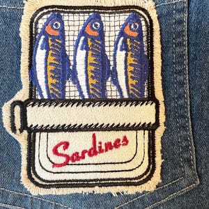 May include: Embroidered patch showing three stylized sardines in a can. The sardines are blue, yellow, and white, set against a white background with a black grid. The word "Sardines" is embroidered in red.