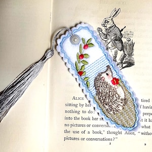 May include: Embroidered bookmark with a hedgehog design, red flower, and silver tassel. The bookmark has a blue scalloped edge and a floral design. The background shows a vintage illustration of a rabbit.