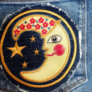 May include: Embroidered patch depicting a crescent moon face with red floral details and gold stars on a dark blue backdrop. The patch has a gold border and a frayed edge, ideal for customising clothing or bags.