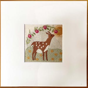 May include: Embroidered artwork of a deer with white spots, on a green grassy hill. Above the deer is a floral arch with pink flowers and a yellow sun. The artwork is framed in a white mount and a wooden frame.