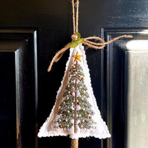 May include: A handcrafted Christmas tree decoration. The white fabric tree is adorned with embroidered details, including a gold star, colourful beads, and a brown trunk. It hangs from a twine loop with a green leaf and a small bell.