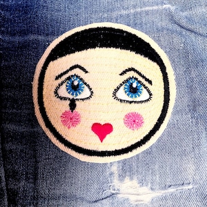 May include: A white embroidered patch with a black outline, depicting a cartoon face with blue eyes, pink cheeks, and a red heart. The face has a black line down the nose.
