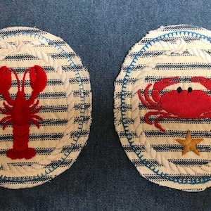 May include: Two round nautical coasters. One features a red lobster, the other a red crab with a small gold starfish. Both have a white and blue striped background, a rope-like border, and blue stitching.