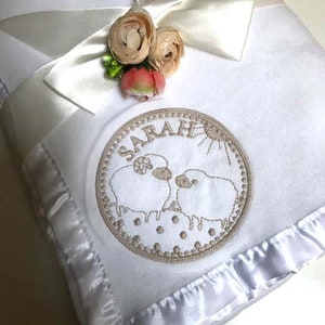 May include: A white baby blanket featuring a satin ruffle trim and a white satin ribbon tied in a bow. The blanket is decorated with a circular embroidery design showing two sheep, the name SARAH, and a sun, accented by three fabric flowers.
