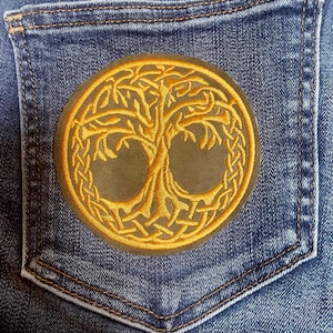 May include: A circular embroidered patch featuring a golden yellow tree of life design with intricate Celtic knotwork borders. The patch is centred on the back pocket of a pair of blue denim jeans with visible stitching and fabric texture.