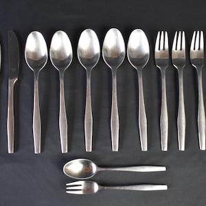 May include: A set of stainless steel flatware, including knives, spoons, and forks, arranged on a dark surface. The spoons and forks have a rounded bowl and tines, respectively. The knives have a simple, pointed blade. The set includes two additional pieces.