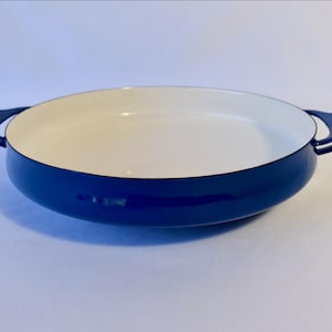 May include: A round, shallow cooking pan with a glossy dark blue exterior and a smooth, cream-colored interior. The pan features two integrated, curved handles on opposite sides, providing a secure grip for lifting and maneuvering the vessel.