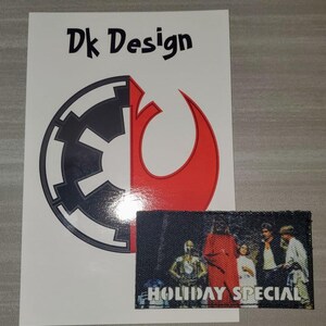 May include: A white sticker with a black and red logo that says "Dk Design". The logo is a combination of the Star Wars Imperial symbol and the Rebel Alliance symbol. Below the sticker is a black patch with a scene from the Star Wars Holiday Special and the text "HOLIDAY SPECIAL".