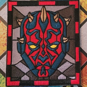 May include: A red, blue, and yellow embroidered patch of Darth Maul's face in a stained glass style. The patch is surrounded by a black border with a red frame.