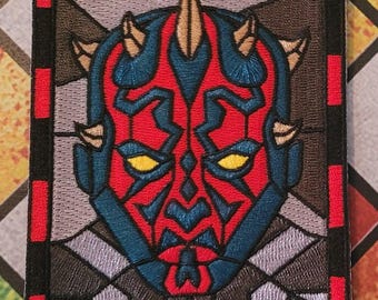 Può includere: Una toppa ricamata con il volto di Darth Maul in stile vetrata. La toppa è rossa, blu e gialla, ed è circondata da un bordo nero con una cornice rossa.