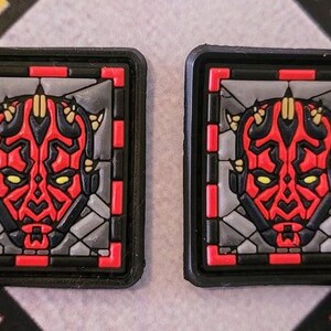 May include: Two square patches featuring a red and black character design with yellow accents, set against a gray background. The design is framed by a black border with red corner accents. The patches are likely for clothing or accessories.
