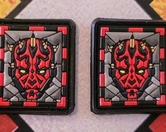 Può includere: Due toppe quadrate con un design di personaggio rosso e nero con accenti gialli, su sfondo grigio. Il design è incorniciato da un bordo nero con accenti rossi agli angoli. Queste toppe sono probabilmente per abbigliamento o accessori.