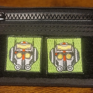 Può includere: Una pochette nera con cerniera a zip con due toppe ricamate in verde e giallo di una testa di robot con una striscia rossa sugli occhi.
