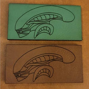 Puede incluir: Dos parches de cuero con un contorno negro de una cabeza alienígena. El parche de arriba es verde y el parche de abajo es marrón.