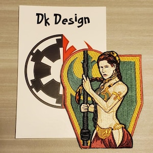 May include: An embroidered patch featuring a woman with long brown hair, wearing a gold and red outfit, holding a staff. The background is green and yellow with a crescent moon symbol. The patch has the text "Dk Design" on it.