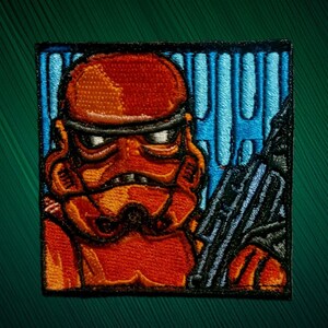May include: A square embroidered patch featuring a stylized orange and red helmeted character with a black visor. The figure is positioned against a bright blue vertical striped background and holds a black weapon in the foreground.