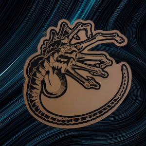 Puede incluir: Una pegatina troquelada marrón y negra con una ilustración estilizada de una criatura con cuerpo segmentado y múltiples extremidades. La pegatina tiene un diseño gráfico detallado y está colocada sobre un fondo azul oscuro con líneas fluidas.