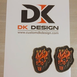 May include: Two black and orange 3D printed patches featuring the face of Darth Maul from Star Wars. The patches are on a white background with the text "DK DESIGN" and "www.customdkdesign.com".