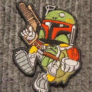 May include: A green, red, and black embroidered patch of a Star Wars character, Boba Fett, holding a blaster rifle. The patch is on a gray knitted fabric.