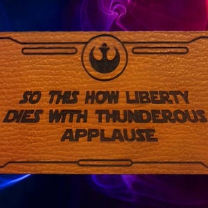 May include: A rectangular orange plaque with a textured surface featuring a black Rebel Alliance symbol at the top center and the black text "SO THIS HOW LIBERTY DIES WITH THUNDEROUS APPLAUSE" engraved in the center, framed by a thin black border.