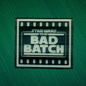May include: A rectangular black and white patch featuring the Star Wars The Bad Batch logo. The design includes a film strip border with white rectangular segments and bold white lettering centered within a frame on a dark background.