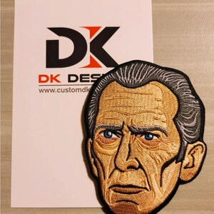 Könnte beinhalten: Gestickter Aufnäher mit dem Gesicht eines Mannes mit einem ernsten Ausdruck. Der Aufnäher ist braun, grau und schwarz. Der Text "DK DESIGNS" und "www.customdk.com" ist im Hintergrund sichtbar.