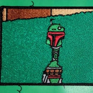 May include: An embroidered patch featuring a green and red Star Wars character, Boba Fett, peeking out from behind a green wall. The patch is surrounded by a black border.