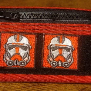 May include: Red and black zippered pouch with two embroidered patches featuring a white and orange helmet design. The pouch has a black zipper and a small black loop on the side.