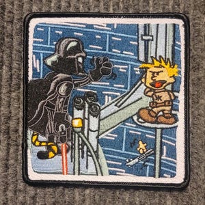 May include: An embroidered patch featuring Darth Vader from Star Wars and a cartoon character resembling Calvin from the comic strip Calvin and Hobbes. Darth Vader is holding a lightsaber and is in a menacing pose. Calvin is standing on a platform and appears to be looking at Darth Vader with a surprised expression.