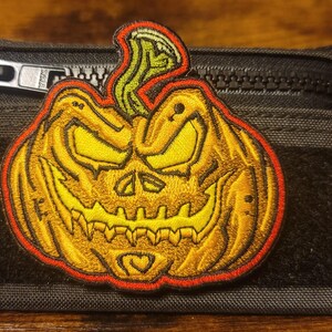 May include: A black zippered pouch with a yellow and orange embroidered pumpkin patch. The pumpkin has a scary face with a wide grin and yellow eyes.