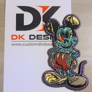 May include: A purple embroidered patch of a cartoon mouse with a skull-like face, red shorts, and yellow shoes. The mouse has a blue and green body with a cracked, skeletal appearance. The patch is on a white background with the text "DK DESI" and "www.customdkdesign" below the patch.