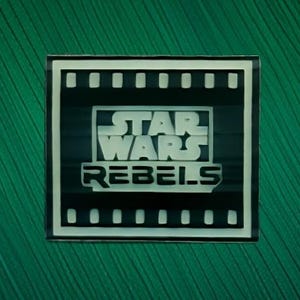 May include: A square-shaped enamel pin featuring a black and white film strip border. The center displays the Star Wars Rebels logo in white text against a black background. The design is clean, with bold lettering and a high-contrast graphic style.
