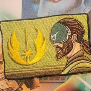 Puede incluir: Un parche bordado amarillo con un sable de luz estilizado y alas, y un perfil de una persona con una visera verde y barba marrón. El parche está sobre un fondo verde.