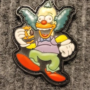 May include: A yellow and blue cartoon clown with a big smile, wearing a purple shirt and a bow tie, holding a brown and yellow burger. The clown is walking with one foot in the air and the other on the ground. The image is a patch with a black border.
