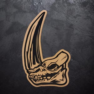 May include: A tan and black sticker depicting a stylized rhino skull with a prominent horn. The design has a detailed, carved appearance, with black lines defining the skull's features and the horn's shape. The sticker is set against a dark, textured background.
