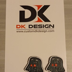 Może przedstawiać: Dwa czarne i białe szpilki emaliowane z postacią Dartha Vadera z czerwonymi oczami. Szpilki znajdują się na białej kartce z tekstem "DK DESIGN" i "www.customdkdesign.com".