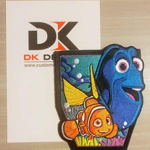 May include: An embroidered patch featuring Dory and Marlin from the movie Finding Nemo. Dory is a blue tang fish with a yellow tail fin and Marlin is an orange clownfish. The patch is surrounded by a black border and has a blue, green, and yellow background.