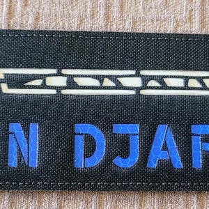 May include: A black fabric patch with a white stylized image of a weapon and the text "DIN DJARIN" in blue.